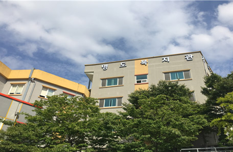 나무위로 보이는 명도복지관 건물전경으로 회색빛 사각건물에 건물 윗쪽에 명도복지관 간판이 달려있는 모습