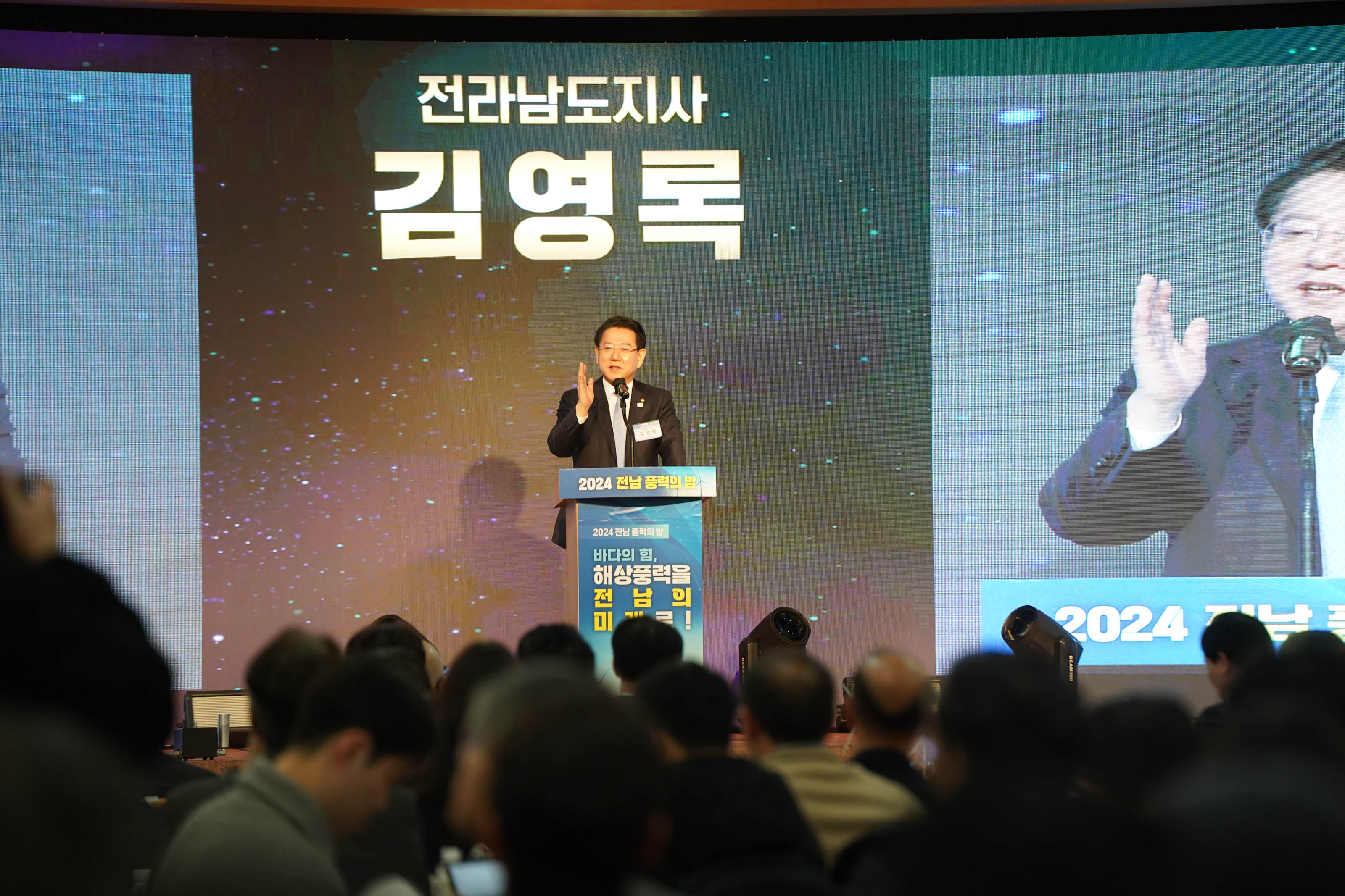 '전라남도지사 김영록'이라는 문구가 적힌 대형 스크린 앞 단상에서 김영록 도지사가 마이크 앞에 서서 손짓을 하며 연설하고 있는 모습. 단상 앞에는 '2024 전남 풍력의 밤'이라는 행사명이 적혀 있고, 무대 아래에는 청중들이 빼곡히 앉아 있는 모습이 보인다.