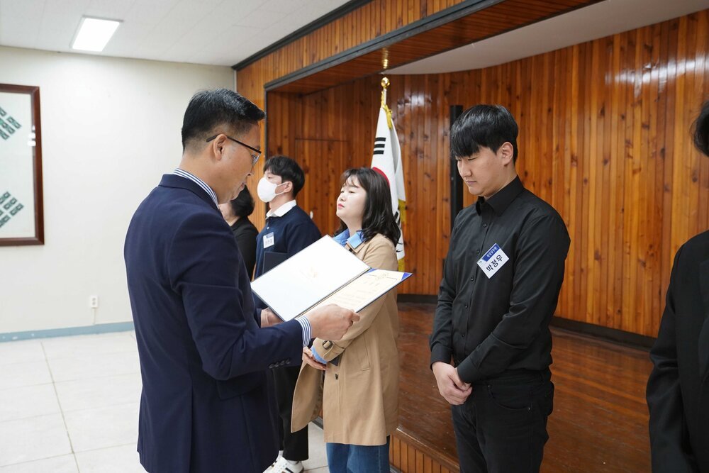 4월 정례조회에 정장을 입고 안경을 낀 남성이 검정색 셔츠를 입은 박정우에게 패를 수여하고 있다